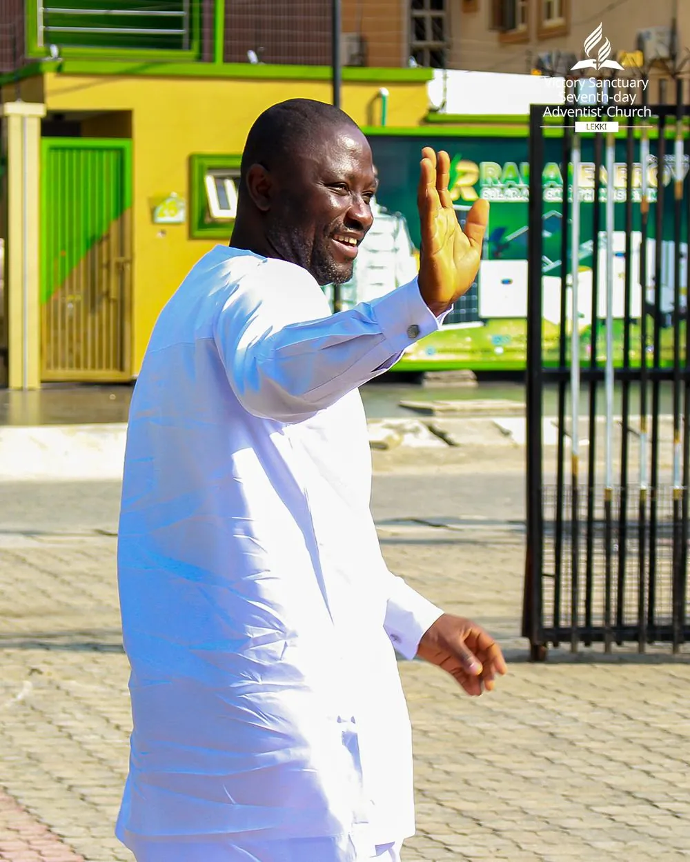 A smiling member in white waving hello at the church gate in Lekki