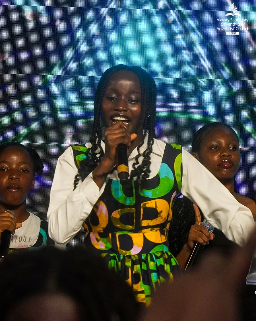 A girl in a green and gold print dress singing on stage at Children’s Day