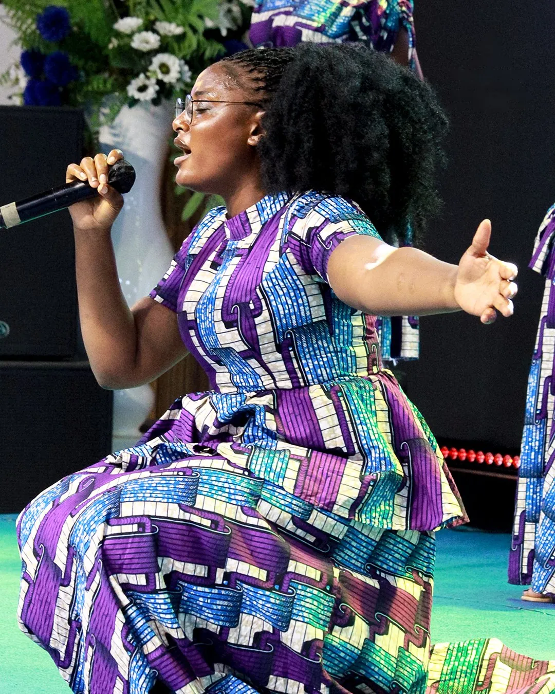 A music minister in a purple print dress singing with joy on stage