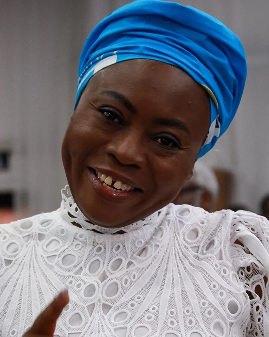 A smiling woman in a blue head wrap and white lace
