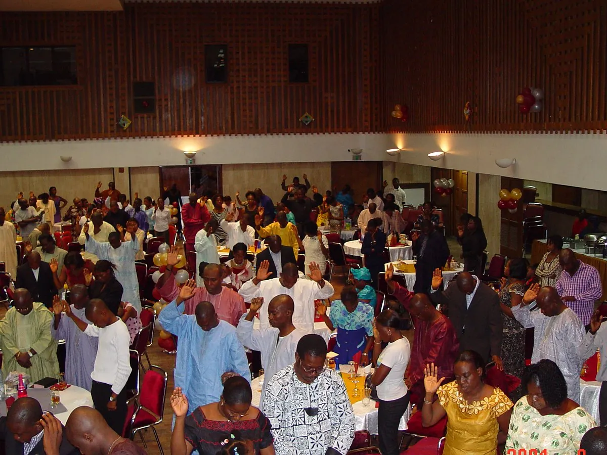 A hall full of round tables, people standing with hands raised in prayer